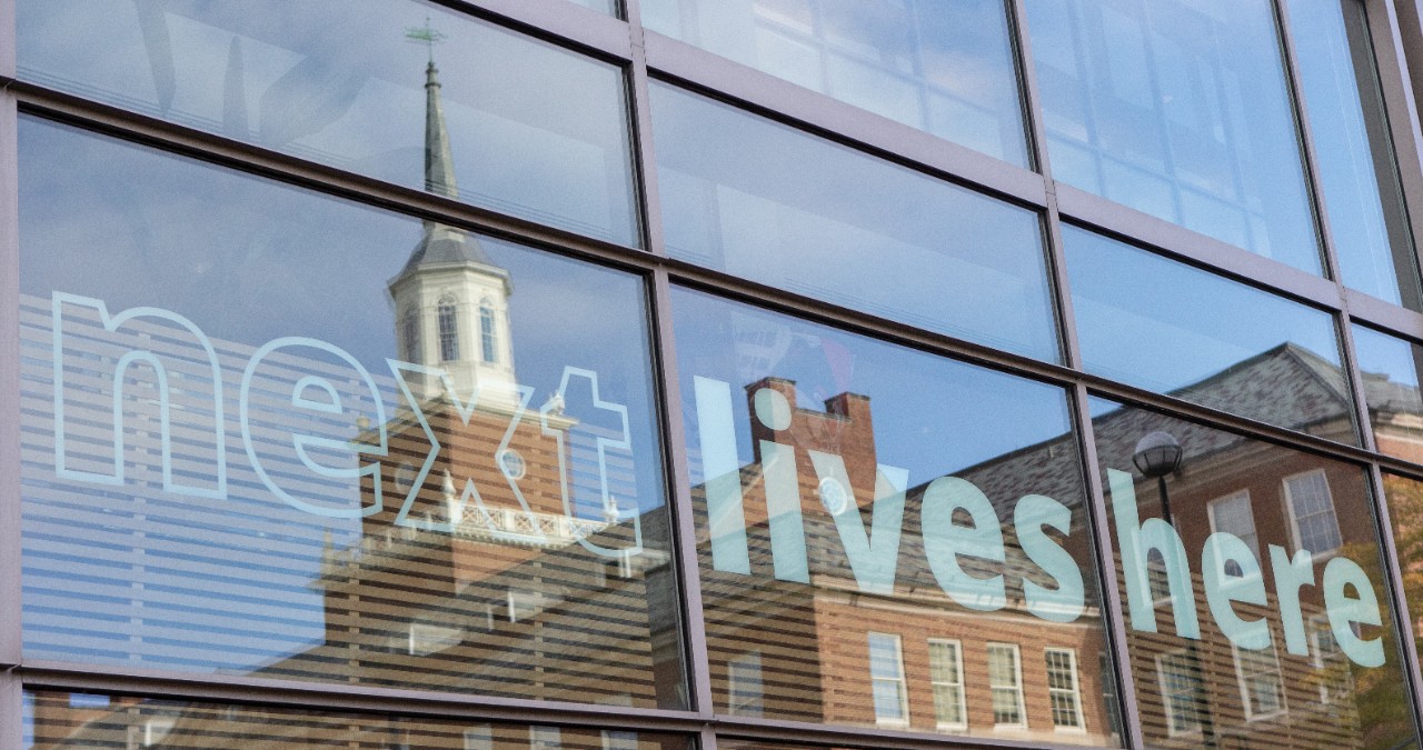 Window reflection showing McMicken Hall and the words Next Lives Here displayed.